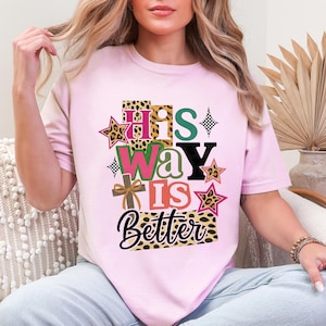 May include: A pale pink t-shirt featuring the text "His Way Is Better" in a layered design with leopard print, stars, and a cross. The design uses a variety of colours, creating a vibrant and faith-based graphic.