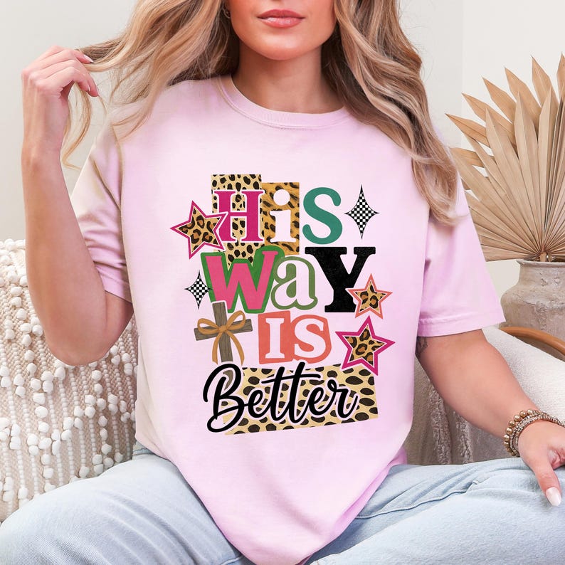 May include: A pale pink t-shirt featuring the text "His Way Is Better" in a layered design with leopard print, stars, and a cross. The design uses a variety of colours, creating a vibrant and faith-based graphic.