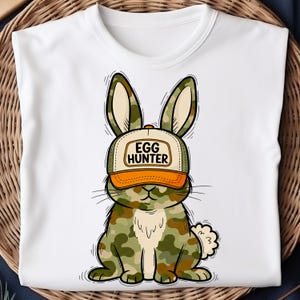 Puede incluir: Camiseta blanca con un conejo de dibujos animados que lleva una gorra de camuflaje. La gorra dice "EGG HUNTER". El conejo tiene un patr&oacute;n de camuflaje en tonos verdes, marrones y beige. El dise&ntilde;o est&aacute; centrado en la camiseta.