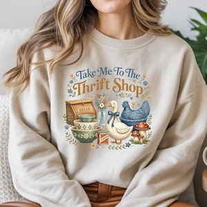 May include: Beige sweatshirt with a graphic that says "Take Me To The Thrift Shop." The graphic features a bread box, bowls, a duck with a blue bow, a blue chicken, mushrooms, and flowers.