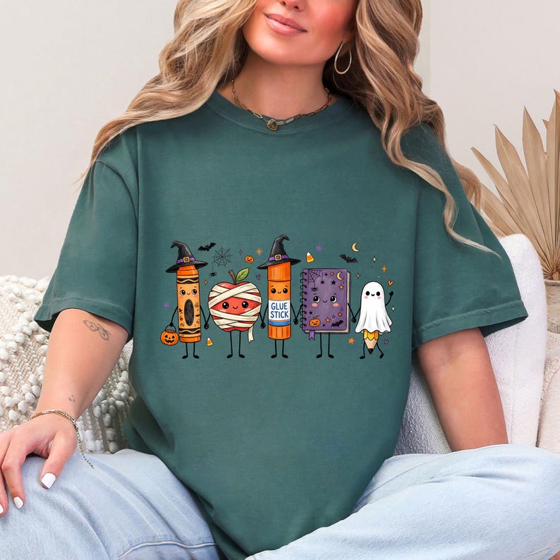 May include: A dark green short-sleeve t-shirt featuring a graphic print of five cute Halloween-themed school supplies holding hands: a crayon in a witch hat, a mummy apple, a glue stick in a witch hat, a purple notebook, and a ghost pencil.