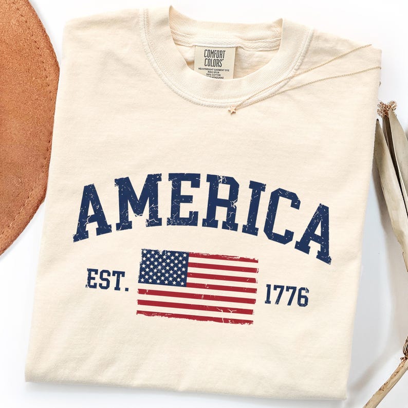 May include: A cream-coloured t-shirt featuring the word AMERICA in a distressed navy blue font arched above a graphic of the United States flag. The text EST. 1776 is printed in navy blue on either side of the flag. The shirt has a Comfort Colors label.