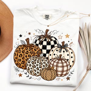May include: A white t-shirt featuring a graphic print of five pumpkins with various patterns including leopard print, black and white checkered, polka dots, and plaid. The design includes small star accents and vine illustrations. The shirt has a Comfort Colors label.