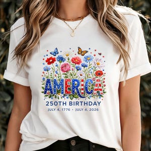K&ouml;nnte beinhalten: Wei&szlig;es T-Shirt mit floralem Design und dem Wort "AMERICA" in Rot, Wei&szlig; und Blau. Das Design umfasst Blumen, Schmetterlinge und den Text "250TH BIRTHDAY JULY 4, 1776 &bull; JULY 4, 2026".