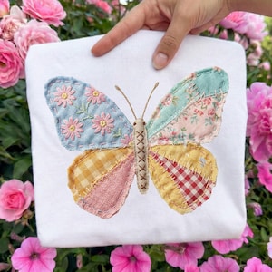 May include: White square fabric with a large, colourful butterfly appliqué. The butterfly wings are patchwork, featuring floral, gingham, and plaid patterns in pastel shades of blue, pink, and yellow. The butterfly's body is beige with stitched details.