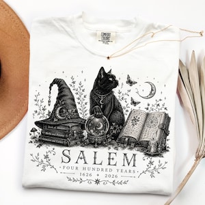 May include: A white t-shirt featuring a black ink illustration of a cat, a witch hat on a stack of books, a crystal ball, and an open book. The text reads SALEM FOUR HUNDRED YEARS 1626 2026. The shirt has a Comfort Colors brand label at the neck.