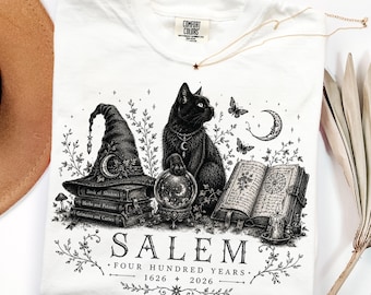 May include: A white t-shirt featuring a black ink illustration of a cat, a witch hat on a stack of books, a crystal ball, and an open book. The text reads SALEM FOUR HUNDRED YEARS 1626 2026. The shirt has a Comfort Colors brand label at the neck.