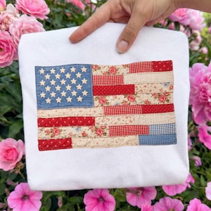 May include: White sweatshirt featuring a patchwork American flag design. The flag incorporates blue denim with white stars, red gingham, floral, and striped fabric patterns. The sweatshirt is held against a backdrop of pink flowers.