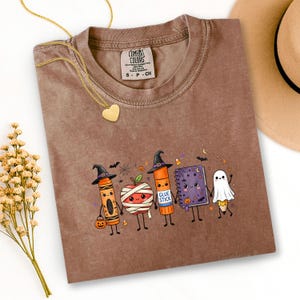 May include: A brown short-sleeve t-shirt featuring a graphic of five cartoon characters: a crayon in a witch hat, a mummy apple, a glue stick in a witch hat, a purple notebook, and a pencil dressed as a ghost, all holding hands with small bats.