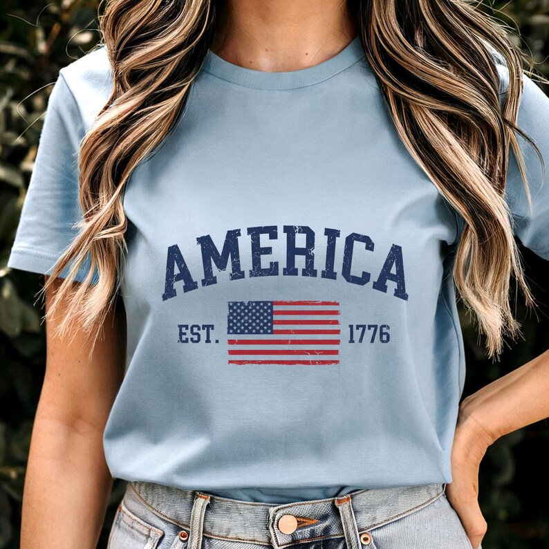 May include: A light blue short-sleeve t-shirt featuring the dark blue text AMERICA in an arched font above a graphic of the United States flag. The text EST. 1776 is printed in dark blue on either side of the flag, all with a distressed finish.