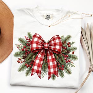 May include: A white short-sleeve t-shirt featuring a graphic of a large red and white gingham bow centered over a cluster of green pine branches, red berries, and small brown pinecones. The inner neck label displays the text Comfort Colors.