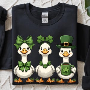 May include: Black sweatshirt featuring three cartoon geese. One goose wears a green bow, another a shamrock headband, and the third a green hat and holds a mug with a shamrock. The geese stand on green grass.