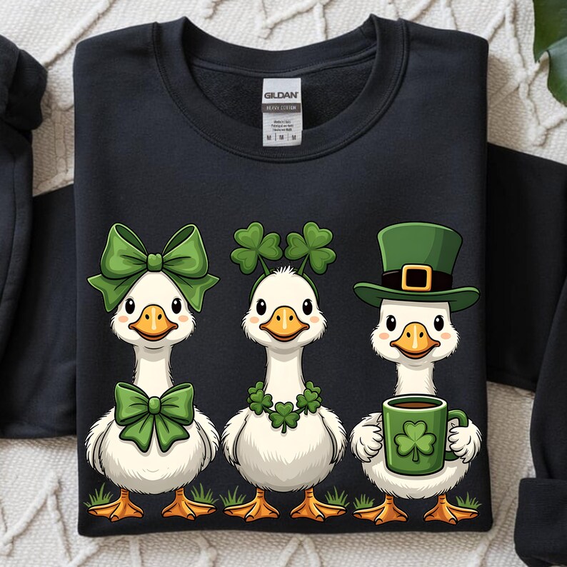 May include: Black sweatshirt featuring three cartoon geese. One goose wears a green bow, another a shamrock headband, and the third a green hat and holds a mug with a shamrock. The geese stand on green grass.