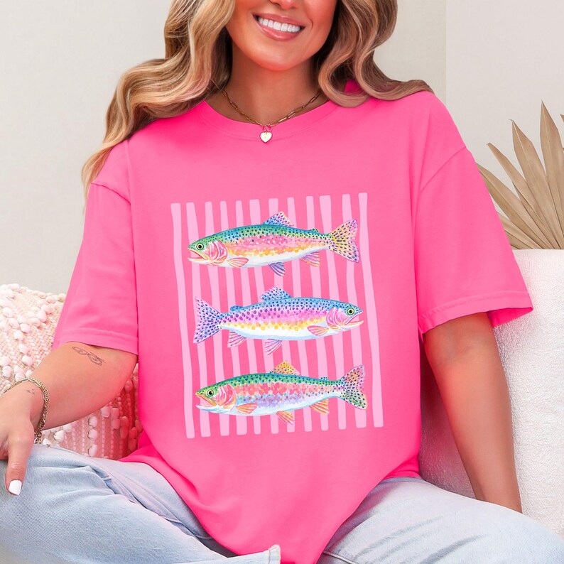 May include: A bright pink t-shirt featuring a graphic of three colorful fish. The fish are depicted in shades of blue, green, yellow, and pink, set against a background of vertical pink stripes. The shirt is a casual, short-sleeved style.