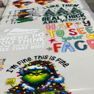 May include: A sheet of printed designs featuring various graphics and text. Designs include colorful birds, Christmas-themed text, and a Grinch character with the text "I'm Fine. This is Fine."
