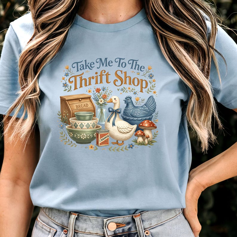 May include: Light blue t-shirt with the text "Take Me To The Thrift Shop." The graphic features a goose with a blue bow, a blue hen, mushrooms, bowls, and a bread box. The design has a vintage, farmhouse aesthetic.