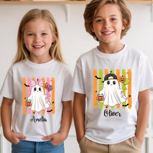 May include: Two white t-shirts featuring Halloween ghost graphics. The left shirt shows a ghost with a pink plaid bow and the name Amelia. The right shirt shows a ghost with a black baseball cap and the name Oliver. Both have orange and green stripes.