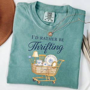 Op de afbeelding: Teal Comfort Colors T-shirt met de tekst "I'd Rather Be Thrifting". De afbeelding toont een winkelwagentje gevuld met vintage porselein, een lamp en een stoel. Een zilveren ketting met een hanger ligt over het shirt.