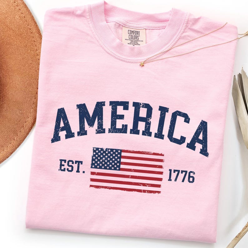 May include: A light pink t-shirt featuring the word AMERICA in dark blue distressed lettering. Below the text is a graphic of the American flag with the text EST. on the left and 1776 on the right. The shirt has a Comfort Colors brand neck label.
