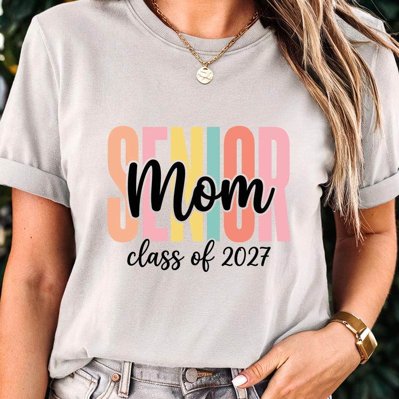 May include: A light gray short-sleeved t-shirt featuring the text "SENIOR" in large, vertical, pastel-colored block letters behind the word "Mom" in black cursive script, with the text "class of 2027" printed in black below it.