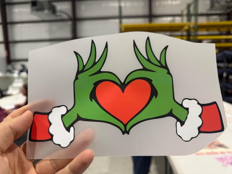 May include: A green hand-shaped heart design with a red heart in the centre. The hands have green fingers and red cuffs with white trim. The design is on a clear, rectangular sheet.