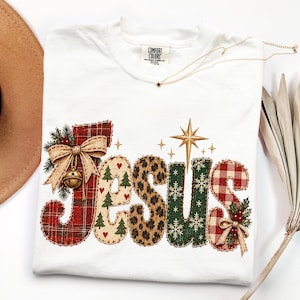 May include: A white t-shirt featuring the word JESUS in large, stitched-style letters. Each letter has a unique pattern including red plaid, cream with green trees, leopard print, dark green with snowflakes, and red gingham with holly accents.