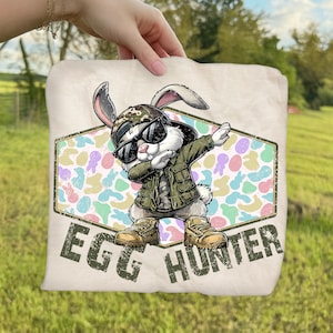 May include: A beige Easter-themed throw pillow featuring a cartoon rabbit wearing sunglasses, a camouflage hat, and a green jacket, dabbing. The pillow has the words "EGG HUNTER" printed on it.