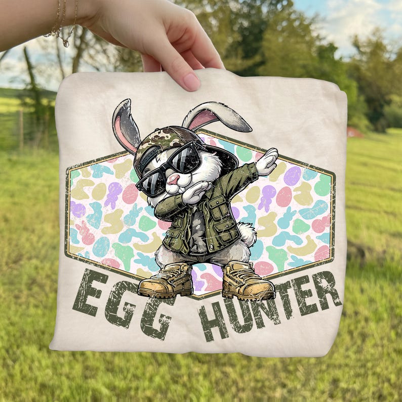May include: A beige Easter-themed throw pillow featuring a cartoon rabbit wearing sunglasses, a camouflage hat, and a green jacket, dabbing. The pillow has the words "EGG HUNTER" printed on it.