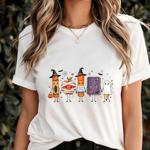 May include: A white t-shirt featuring a graphic print of five cartoon school supplies dressed for Halloween: an orange crayon in a witch hat, a mummy apple, an orange glue stick in a witch hat, a purple notebook, and a pencil dressed as a ghost.