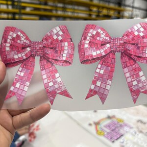 May include: Two pink bow-shaped stickers with a mosaic tile design. The bows have long, pointed tails and are in a vibrant shade of pink with white accents. The background is a light, translucent material.