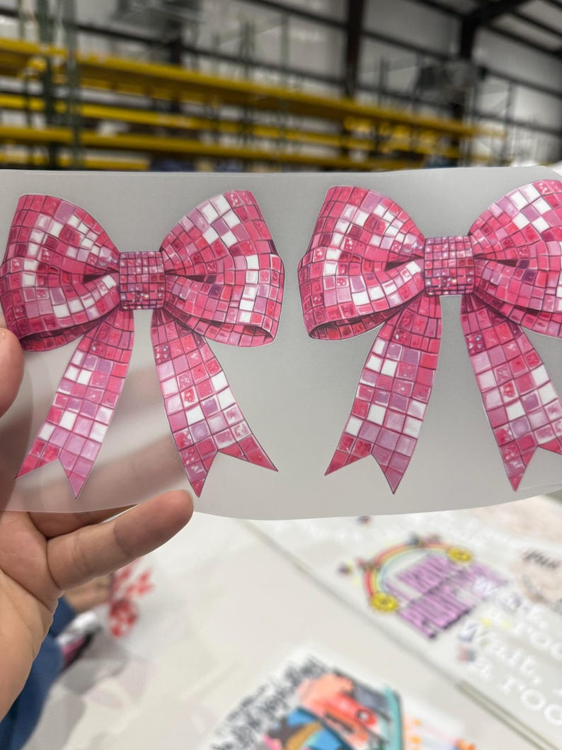 May include: Two pink bow-shaped stickers with a mosaic tile design. The bows have long, pointed tails and are in a vibrant shade of pink with white accents. The background is a light, translucent material.
