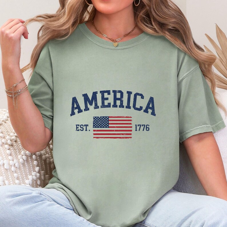 May include: A sage green short-sleeve t-shirt featuring the dark blue text AMERICA in an arched font above a graphic of the United States flag. The text EST. and 1776 are printed in dark blue on either side of the flag graphic on the chest.