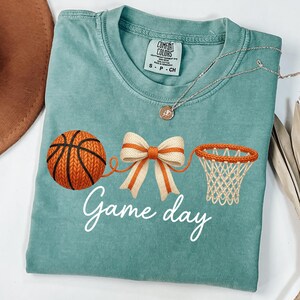 May include: A teal Comfort Colors t-shirt featuring a basketball, a bow, and a basketball net graphic. The words "Game day" are written in white script. A silver necklace with a pendant is draped across the shirt.