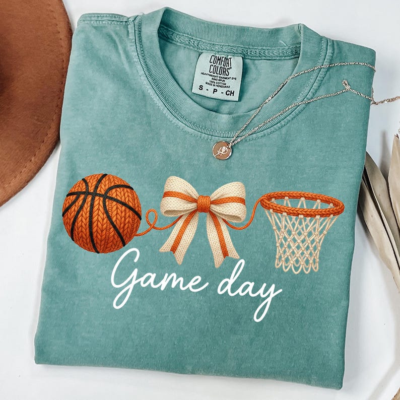 May include: A teal Comfort Colors t-shirt featuring a basketball, a bow, and a basketball net graphic. The words "Game day" are written in white script. A silver necklace with a pendant is draped across the shirt.