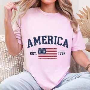 May include: A light pink short-sleeve t-shirt featuring the dark blue text AMERICA arched over a distressed graphic of the United States flag. The text EST. and 1776 are printed in dark blue on either side of the flag graphic on the front.