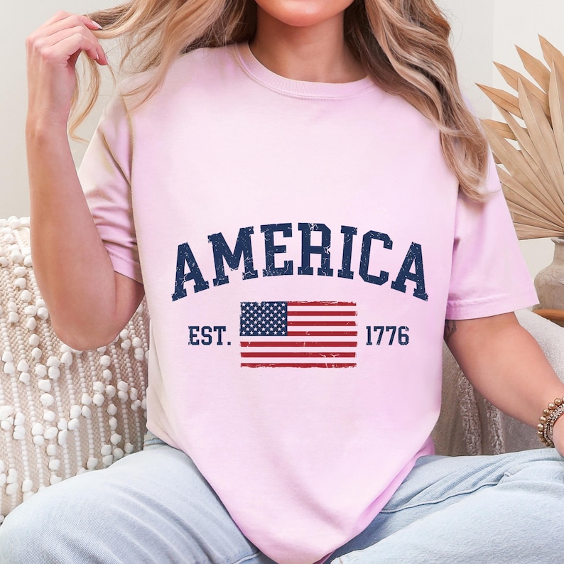 May include: A light pink short-sleeve t-shirt featuring the dark blue text AMERICA arched over a distressed graphic of the United States flag. The text EST. and 1776 are printed in dark blue on either side of the flag graphic on the front.