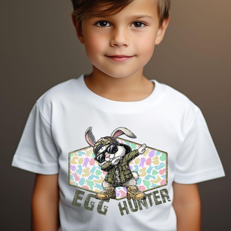 May include: White t-shirt featuring a cartoon rabbit wearing sunglasses, a camouflage hat, and a green jacket, striking a dabbing pose. The shirt has the text "EGG HUNTER" in a distressed font, perfect for Easter.