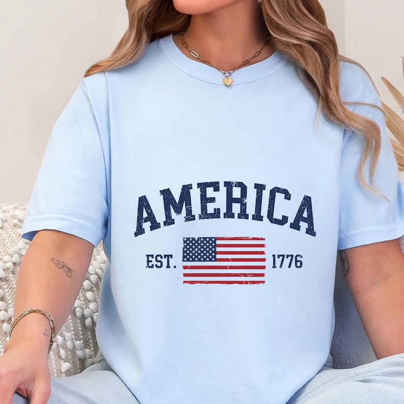 May include: A light blue short-sleeve t-shirt featuring the dark blue text AMERICA arched above a graphic of the United States flag. Below the flag, the text EST. 1776 is printed in a dark blue, distressed, vintage-style font.