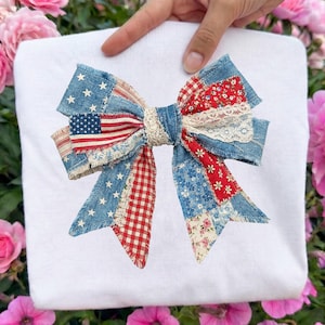 May include: A white t-shirt featuring a large, decorative bow. The bow is made of patchwork denim, red gingham, floral, and lace fabrics. It incorporates an American flag design with stars and stripes, creating a patriotic theme.