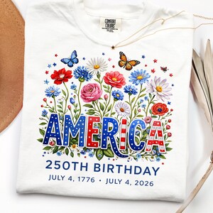 K&ouml;nnte beinhalten: Wei&szlig;es T-Shirt mit floralem Design und dem Wort "AMERICA" in Rot, Wei&szlig; und Blau. Das Shirt zeigt Schmetterlinge, Blumen und den Text "250TH BIRTHDAY JULY 4, 1776 &bull; JULY 4, 2026". Eine goldene Halskette mit Sternanh&auml;nger ist ebenfalls zu sehen.