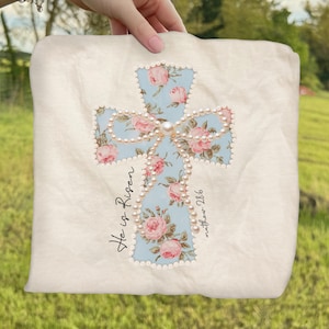 May include: A cream-colored pillow with a light blue cross design. The cross is decorated with pink roses and faux pearls. The words "He is Risen" and "matthew 28:6" are written on the pillow.