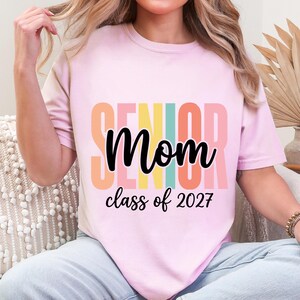 May include: A light pink t-shirt featuring the word SENIOR in large, vertical, multi-colored block letters behind the black cursive text Mom and the black printed text class of 2027. The design is centered on the front of the garment.