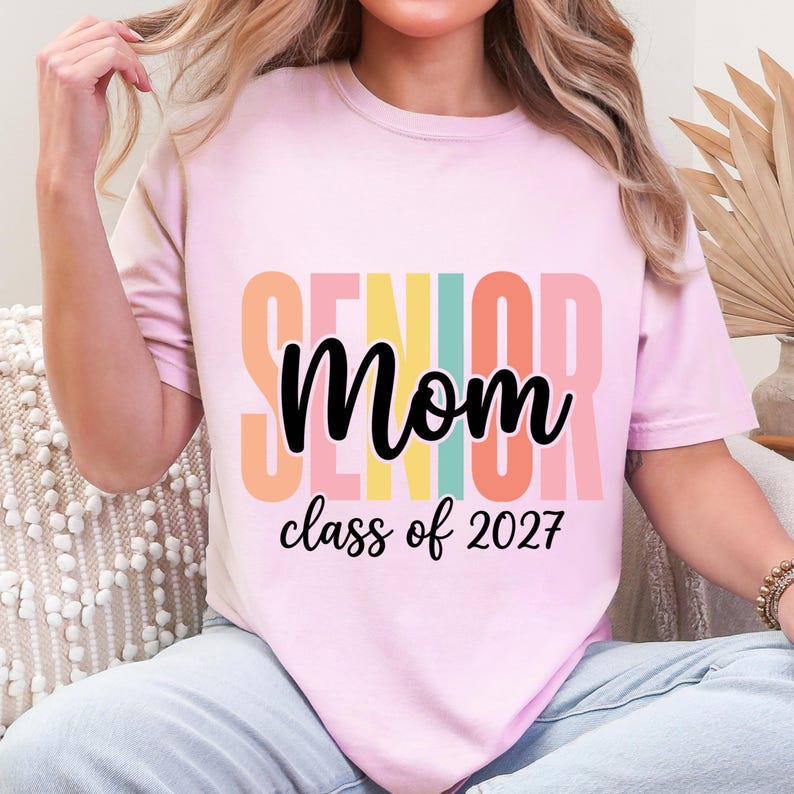May include: A light pink t-shirt featuring the word SENIOR in large, vertical, multi-colored block letters behind the black cursive text Mom and the black printed text class of 2027. The design is centered on the front of the garment.