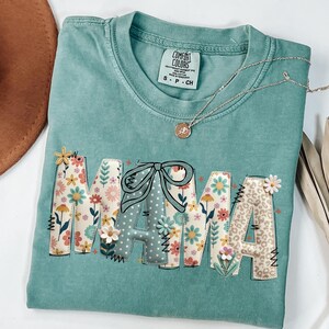 May include: A teal Comfort Colors t-shirt with the word "MAMA" in a floral and patterned design. The letter "M" is decorated with flowers, the "A" has a bow, and the last "A" has a leopard print. A gold necklace with a pendant rests on the shirt.