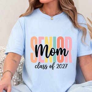 May include: A light blue t-shirt featuring the word SENIOR in large, vertical, multi-colored block letters behind the word Mom in black script. Below this, the text reads class of 2027 in black script. The shirt has a crew neckline.
