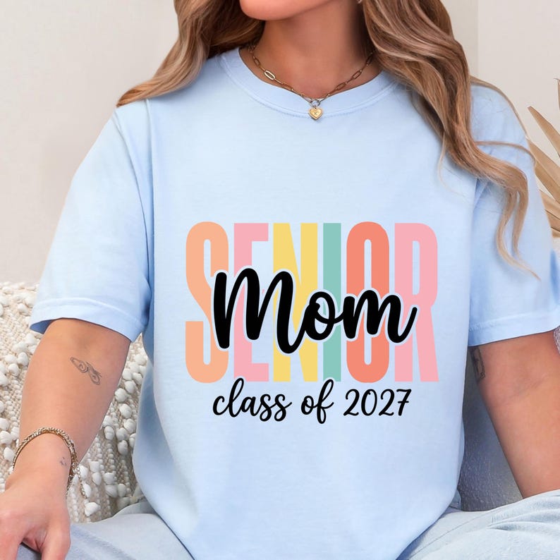 May include: A light blue t-shirt featuring the word SENIOR in large, vertical, multi-colored block letters behind the word Mom in black script. Below this, the text reads class of 2027 in black script. The shirt has a crew neckline.