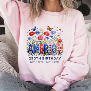 K&ouml;nnte beinhalten: Hellrosa Sweatshirt mit floralem Design und dem Wort "AMERICA" in Rot, Wei&szlig; und Blau. Das Design umfasst Schmetterlinge und Blumen. Der Text "250TH BIRTHDAY" und Daten stehen darunter.