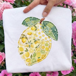 May include: White sweatshirt featuring a large, embroidered lemon design. The lemon is composed of various yellow and green patterned fabrics, with green leaves and a brown stem. The design is held by a hand, set against a background of pink flowers.