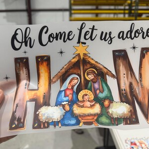 May include: A printed graphic featuring a nativity scene with the text "Oh Come Let us adore HAM." The image depicts Mary, Joseph, baby Jesus, sheep, and a star. The letters "H", "A", and "M" are stylized with a brown and orange gradient.