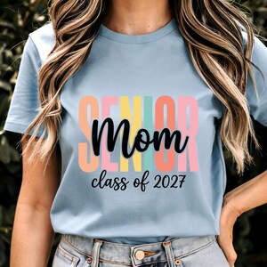 May include: A light blue short-sleeve t-shirt featuring the word SENIOR in large, colorful block letters behind the black cursive text Mom and the black script text class of 2027. The design is centered on the front of the shirt.